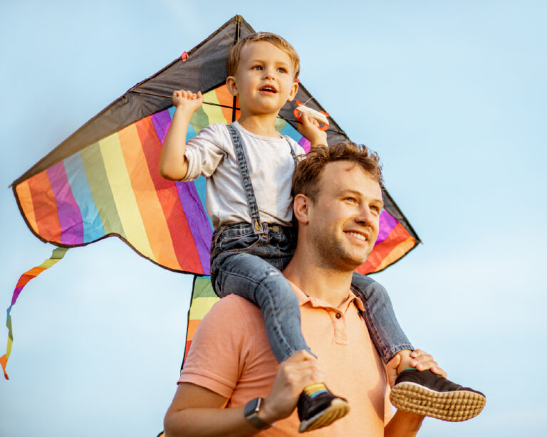 un petit garçon qui tient un cerf-volant coloré dans ses mains. Il est sur les épaules de son père.